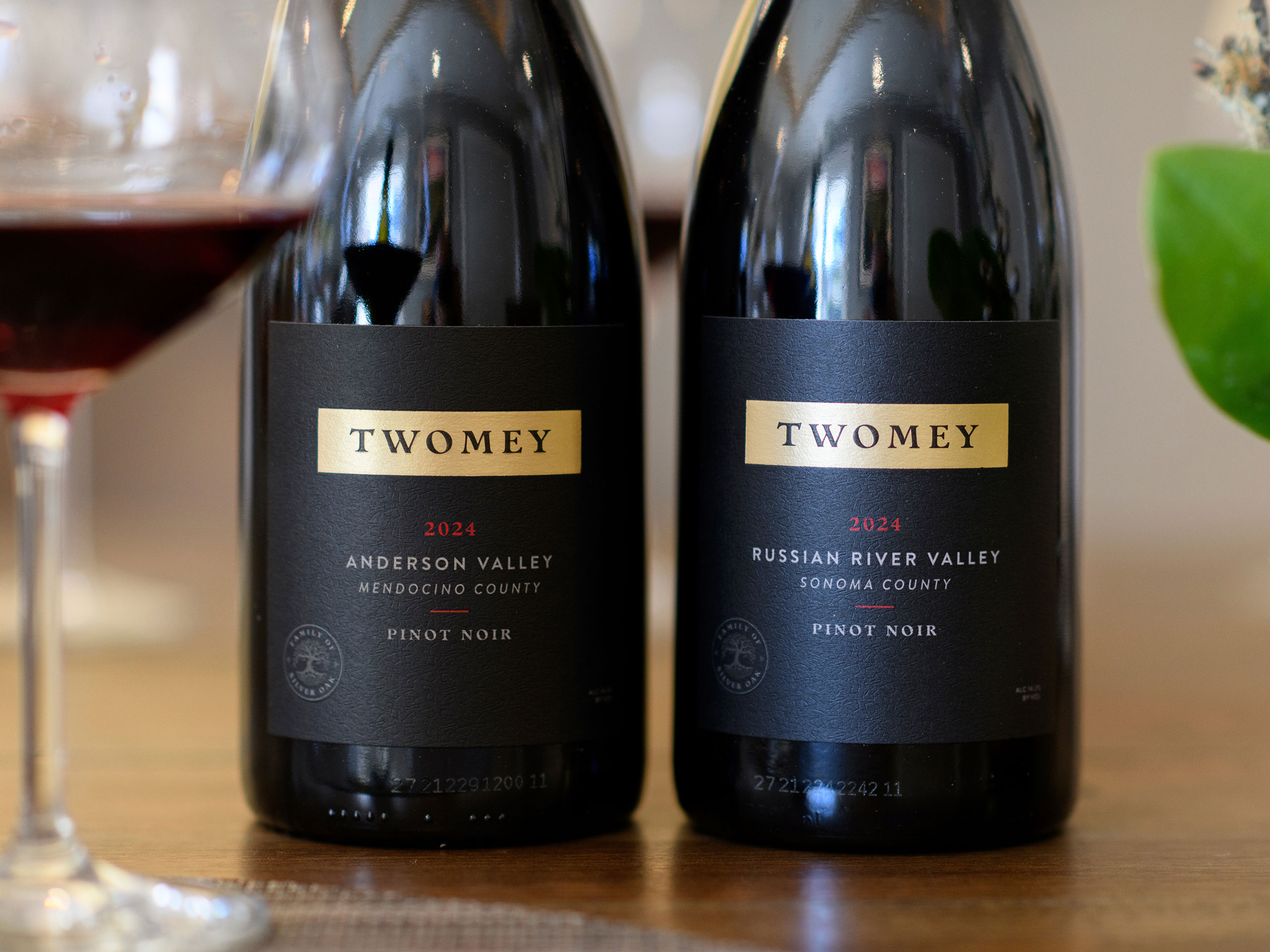 2024 Twomey Anderson Valley and 2024 Twomey Russian River Valley Pinot Noir wine bottles on a wood table with a wine glass